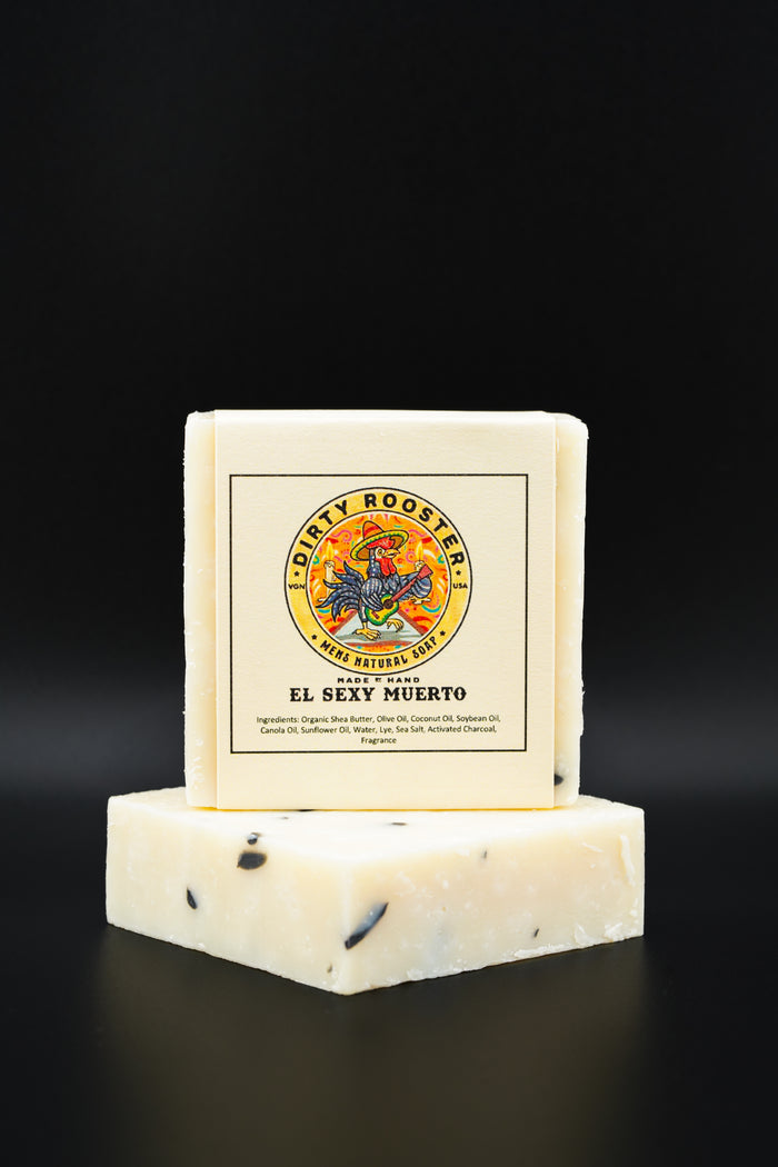 El Sexy Muerto handmade natural soap bar for men, wrapped, standing on an unwrapped cream bar with black flecks