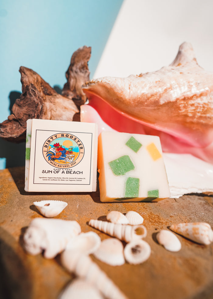 Sun of a Beach handmade natural soap bar for men beside an unwrapped cream bar with green and yellow chunks, with seashells and driftwood
